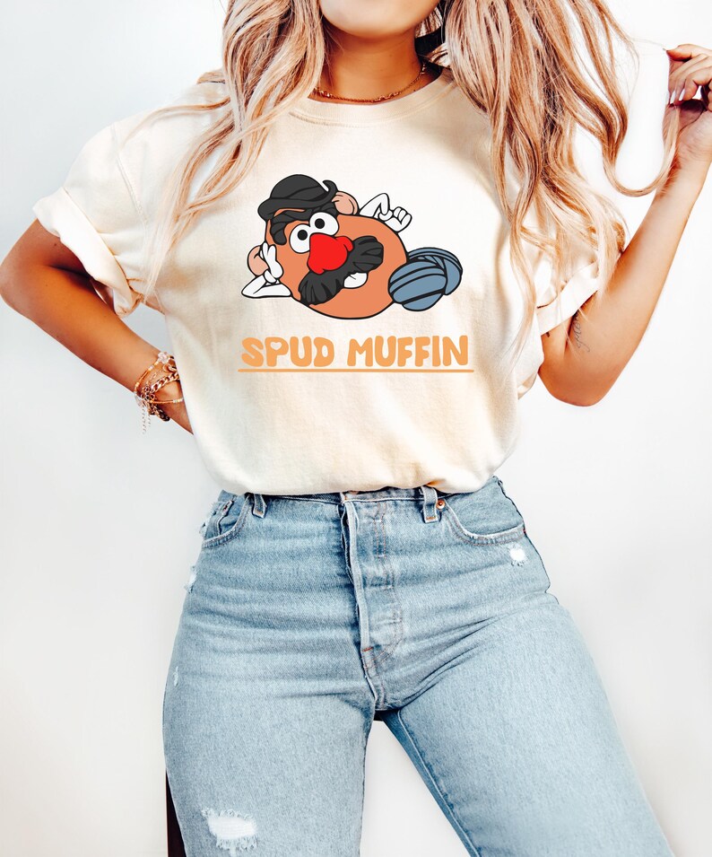 Puede incluir: Camiseta color crema con un personaje de dibujos animados de patata con sombrero negro, bigote y nariz roja. El texto "SPUD MUFFIN" est&aacute; impreso en naranja. La persona lleva vaqueros azul claro.