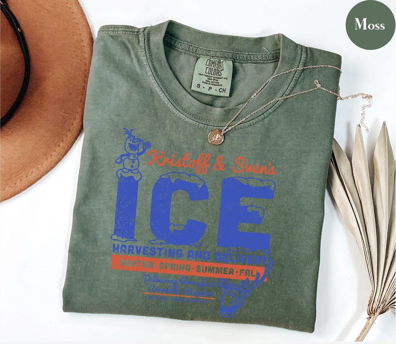 May include: A moss green t-shirt with a graphic that reads "Kristoff & Sven's ICE Harvesting and Delivery". The text is in blue and red. A gold necklace with a pendant is on the shirt.