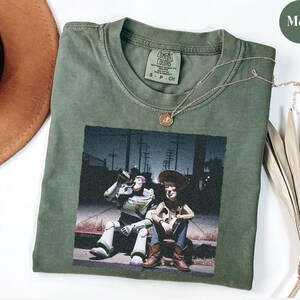 May include: A moss green short-sleeve t-shirt featuring a graphic print of Buzz Lightyear and Woody sitting on a curb at night. The shirt has a Comfort Colors brand label at the inner neckline and is made of cotton. A gold-toned necklace rests on top.