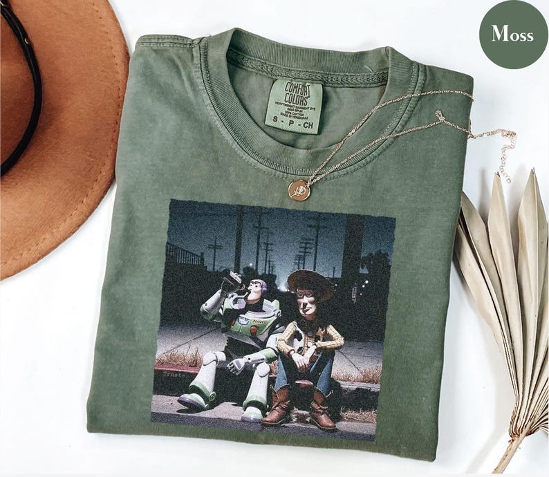 May include: A moss green short-sleeve t-shirt featuring a graphic print of Buzz Lightyear and Woody sitting on a curb at night. The shirt has a Comfort Colors brand label at the inner neckline and is made of cotton. A gold-toned necklace rests on top.