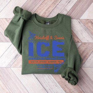 Puede incluir: Una sudadera verde oliva con el texto "Kristoff & Sven's ICE" en naranja y azul. Debajo est&aacute;n las palabras "Harvesting Ice" y "Winter, Spring, Summer, Fall". La sudadera est&aacute; doblada sobre una superficie de madera marr&oacute;n claro.