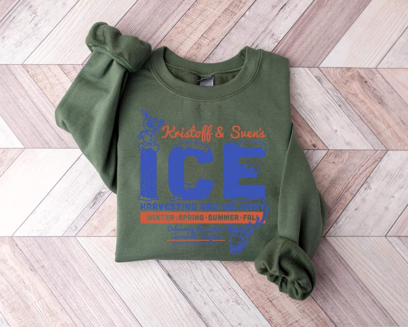 Puede incluir: Una sudadera verde oliva con el texto "Kristoff & Sven's ICE" en naranja y azul. Debajo est&aacute;n las palabras "Harvesting Ice" y "Winter, Spring, Summer, Fall". La sudadera est&aacute; doblada sobre una superficie de madera marr&oacute;n claro.