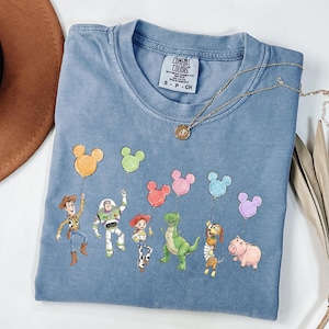 May include: A blue short-sleeved t-shirt featuring a graphic print of six animated characters holding colourful Mickey Mouse-shaped balloons. The shirt has a crew neckline and a white interior label with the text Comfort Colors S P CH.