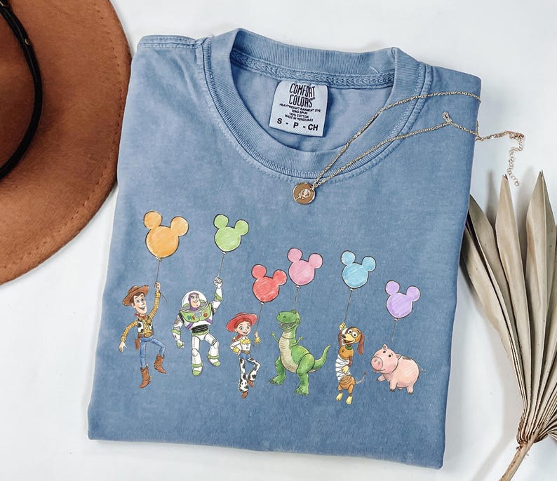 May include: A blue short-sleeved t-shirt featuring a graphic print of six animated characters holding colourful Mickey Mouse-shaped balloons. The shirt has a crew neckline and a white interior label with the text Comfort Colors S P CH.