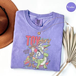 K&ouml;nnte beinhalten: Ein lila Rundhals-T-Shirt mit einer Toy Story-Grafik mit Woody, Buzz Lightyear und Rex. Der Text "Toy Story" und "Since 1995" sind ebenfalls abgebildet. Eine goldene Halskette und ein brauner Hut sind ebenfalls sichtbar.