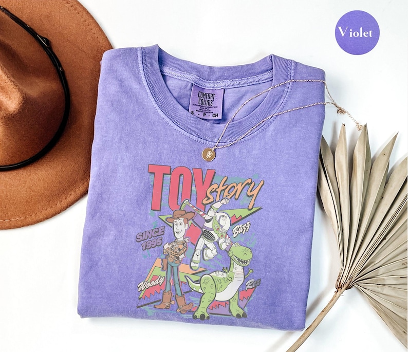 K&ouml;nnte beinhalten: Ein lila Rundhals-T-Shirt mit einer Toy Story-Grafik mit Woody, Buzz Lightyear und Rex. Der Text "Toy Story" und "Since 1995" sind ebenfalls abgebildet. Eine goldene Halskette und ein brauner Hut sind ebenfalls sichtbar.
