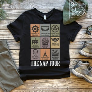 May include: Black t-shirt with a colorful graphic featuring nine squares with various icons and text, including "THE NAP TOUR." The shirt is paired with olive-green pants, hiking boots, and a green bandana. A compass is also visible.