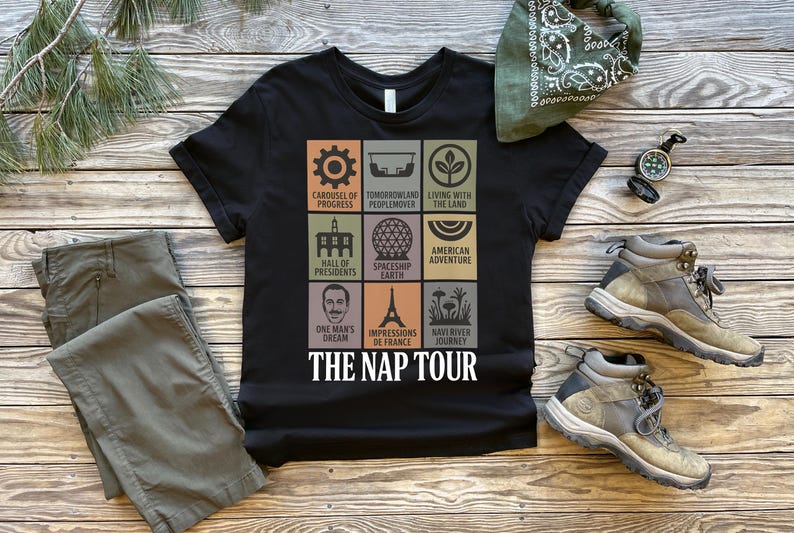 May include: Black t-shirt with a colorful graphic featuring nine squares with various icons and text, including "THE NAP TOUR." The shirt is paired with olive-green pants, hiking boots, and a green bandana. A compass is also visible.