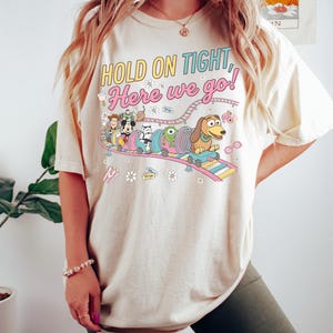 May include: A cream-colored t-shirt featuring a graphic of various cartoon characters riding a roller coaster. The text "HOLD ON TIGHT, Here we go!" is printed above the illustration, which includes small icons of popcorn, a camera, and flowers.