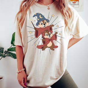 May include: Cream-colored t-shirt featuring a cartoon illustration of two chipmunks dressed as wizards. One chipmunk wears a blue wizard hat with a crescent moon and stars, and holds a wand. The other chipmunk wears a red cloak. Gold stars radiate from the chipmunks.