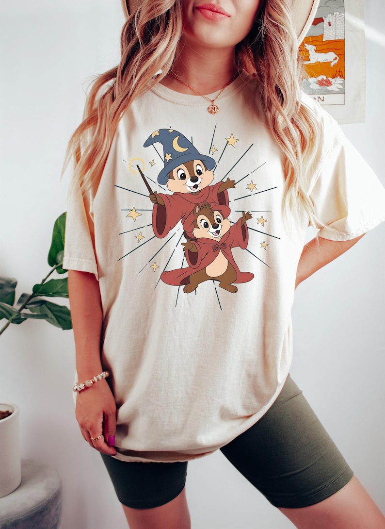 May include: Cream-colored t-shirt featuring a cartoon illustration of two chipmunks dressed as wizards. One chipmunk wears a blue wizard hat with a crescent moon and stars, and holds a wand. The other chipmunk wears a red cloak. Gold stars radiate from the chipmunks.
