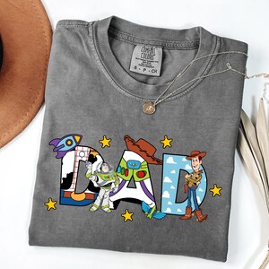 May include: Grey t-shirt featuring the word "DAD" in a Toy Story design. The letters are decorated with images of a rocket, Buzz Lightyear, and Woody. Yellow stars are scattered around the letters. A silver necklace is draped across the shirt.