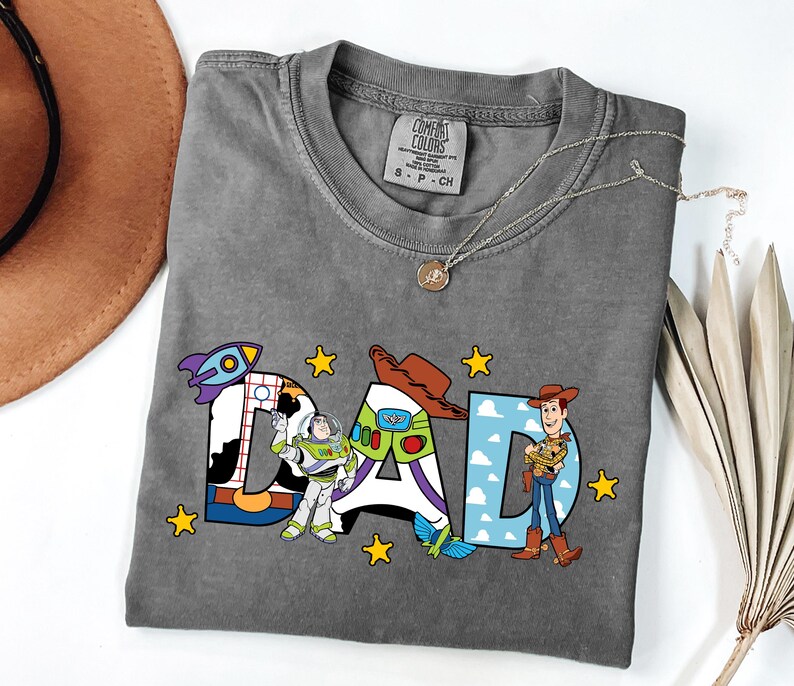 May include: Grey t-shirt featuring the word "DAD" in a Toy Story design. The letters are decorated with images of a rocket, Buzz Lightyear, and Woody. Yellow stars are scattered around the letters. A silver necklace is draped across the shirt.