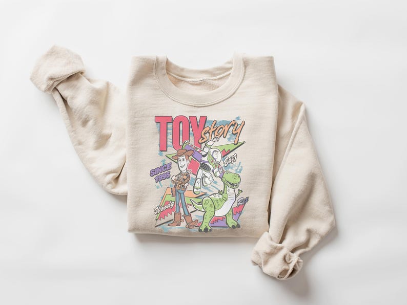 K&ouml;nnte beinhalten: Beiges Sweatshirt mit Rundhalsausschnitt und farbenfroher Grafik von Toy Story-Charakteren. Das Design zeigt Woody, Buzz Lightyear, Rex und den Text "TOY STORY" und "SINCE 1995". Das Sweatshirt ist gefaltet, die &Auml;rmel sind hochgekrempelt.