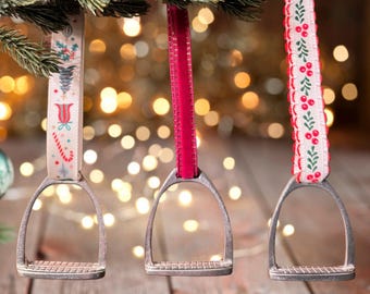 May include: Three silver stirrup ornaments with different patterned ribbons. The ribbons are white with red and green accents. The ornaments are hanging from a Christmas tree.