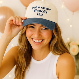 Puede incluir: Gorra de béisbol color crema con visera azul marino. La gorra lleva la frase "put it on my fiancé's tab" en letras azul marino. La gorra la lleva una persona.