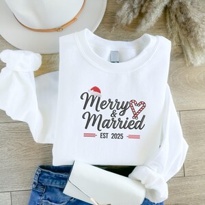 Op de afbeelding: Witte crewneck sweatshirt met de tekst "Merry & Married" in zwart script, een kerstmuts en een zuurstokhart. De tekst "EST 2025" staat eronder in het rood. De sweatshirt is op een houten oppervlak gevouwen.