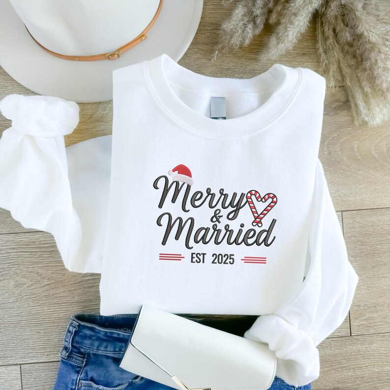 Op de afbeelding: Witte crewneck sweatshirt met de tekst "Merry & Married" in zwart script, een kerstmuts en een zuurstokhart. De tekst "EST 2025" staat eronder in het rood. De sweatshirt is op een houten oppervlak gevouwen.