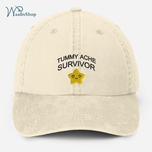 Könnte beinhalten: Eine beige Baumwoll-Baseballkappe mit dem Text "TUMMY ACHESURVIVOR", der in Schwarz gestickt ist. Unter dem Text ist ein gelber Stern mit einem lächelnden Gesicht gestickt.