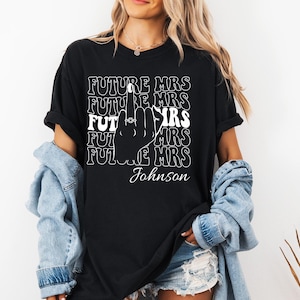 以下が含まれることがあります： 「FUTURE MRS」の文字が重ねてデザインされた黒いTシャツ。指輪をはめた手のシルエットが描かれています。「Johnson」という名前が下にプリントされています。カジュアルなスタイルのシャツです。