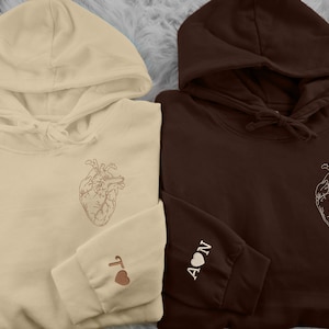 Puede incluir: Dos sudaderas con capucha, una beige y otra marrón, con un gráfico de un corazón en la parte delantera y las palabras "I" y "AON" en las mangas.