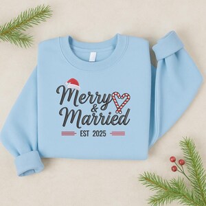 Op de afbeelding: Lichtblauw sweatshirt met de tekst "Merry & Married" in zwart script, een kerstmuts en een zuurstokhart. De tekst "EST 2025" staat eronder. Feestelijke kleding voor koppels.
