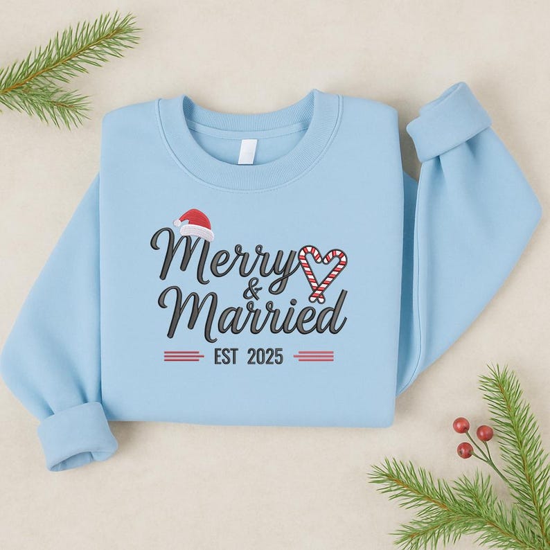 Op de afbeelding: Lichtblauw sweatshirt met de tekst "Merry & Married" in zwart script, een kerstmuts en een zuurstokhart. De tekst "EST 2025" staat eronder. Feestelijke kleding voor koppels.