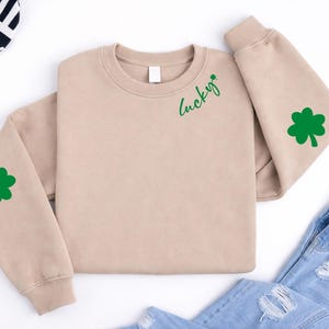Könnte beinhalten: Beiges Sweatshirt mit grünen Akzenten. Das Wort "Lucky" ist in Schreibschrift auf der Brust geschrieben, daneben ein grünes Kleeblatt. Ein grünes Kleeblatt befindet sich auch auf dem linken Ärmel. Hellblaue Jeans im Hintergrund.