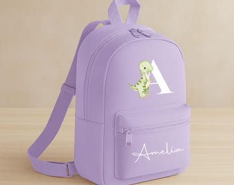 May include: A lavender backpack with a front pocket and adjustable straps. The backpack features a white letter "A" with a green dinosaur illustration and the name "Amelia" in white script.