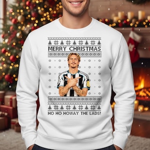Puede incluir: Jersey navideño blanco con un diseño festivo. El jersey presenta el texto "MERRY CHRISTMAS" y "HO HO HOWAY THE LADS!". También incluye un gráfico de una persona con los brazos cruzados. El jersey tiene un patrón de copos de nieve y árboles de Navidad.