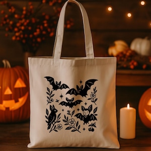 Może przedstawiać: Kremowa torba typu tote z czarnym wzorem nietoperzy i kwiatów. Wzór zawiera nietoperze, kwiaty, gwiazdy i małego ptaka. Tło obejmuje podświetlane lampiony z dyni, świece i dynie, tworząc motyw Halloween.