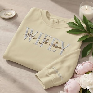 May include: A cream-coloured sweatshirt embroidered with "Mrs. Jackson Wifey" in silver and gold thread. The sleeve cuff displays "01.25.2024". A small dish with rings and a lit candle are also present.