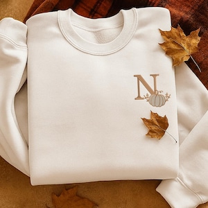 Könnte beinhalten: Cremefarbenes Sweatshirt mit Rundhalsausschnitt, mit einem braunen Buchstaben "N" und einem Kürbisdesign. Zwei getrocknete Herbstblätter sind in der Nähe des Designs platziert. Das Sweatshirt wird auf einer braunen und orangefarbenen karierten Decke präsentiert.