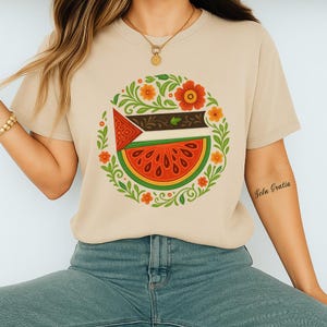 May include: Beige t-shirt with a circular design. The design includes a watermelon slice, a flag, and floral elements in red, orange, green, and brown. The design is centered on the front of the shirt.