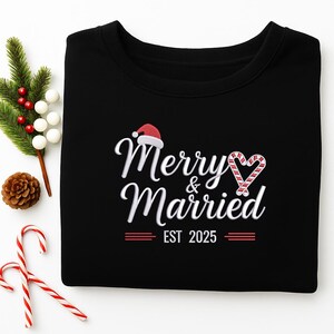Op de afbeelding: Zwart sweatshirt met de tekst "Merry & Married" in witte letters, een kerstmuts en een zuurstokhart. De tekst "EST 2025" staat eronder. Feestelijke vakantiekleding.
