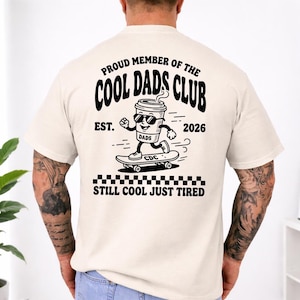 May include: A cream-coloured t-shirt featuring a black graphic of a coffee cup on a skateboard, wearing sunglasses. The text on the shirt reads "PROUD MEMBER OF THE COOL DADS CLUB EST. 2026" and "STILL COOL JUST TIRED".