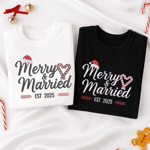 Op de afbeelding: Twee sweatshirts, &eacute;&eacute;n wit en &eacute;&eacute;n zwart, met de tekst "Merry & Married EST 2025". Het ontwerp bevat een kerstmuts en een zuurstokhart. Feestelijke kleding voor een stel.