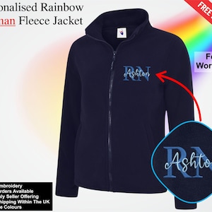 May include: A navy blue fleece jacket with a full zip and a high collar. The jacket features the name "Ashton" embroidered on the chest. The text "Personalised Rainbow Woman Fleece Jacket" is at the top of the image.