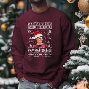 Könnte beinhalten: Ein weinroter Pullover mit einem weißen Weihnachtsmotiv, das eine Person mit einem Weihnachtsmann-Hut und dem Text "GARNACHO HO HO" und "MERRY CHRISTMAS" zeigt.