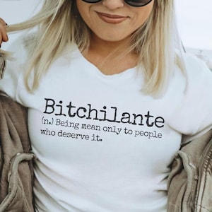 May include: White t-shirt with the word "Bitchilante" printed in black, along with a definition. The person in the image is wearing a gray baseball cap, black sunglasses, and a gray jacket. The person is holding a coffee cup.