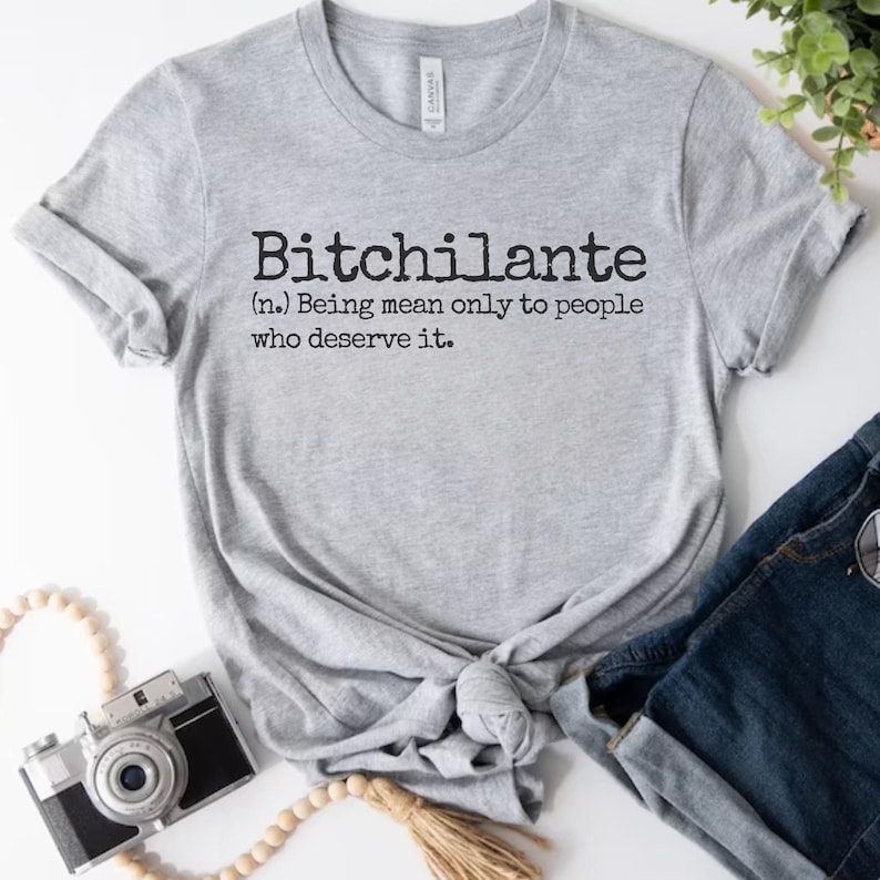 May include: A heather gray t-shirt with the word "Bitchilante" printed in black, along with its definition: "Being mean only to people who deserve it." The shirt is styled with rolled sleeves and a knot at the waist.
