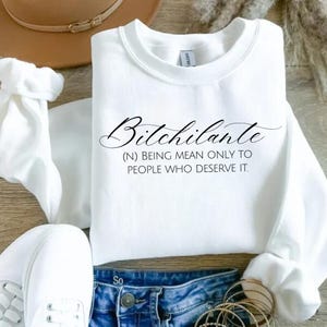 May include: White sweatshirt with the text "Bitchilante (N) BEING MEAN ONLY TO PEOPLE WHO DESERVE IT." The sweatshirt is paired with blue jeans, white sneakers, and gold bracelets. A brown hat is visible in the background.