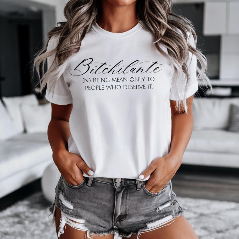 May include: White t-shirt with the word "Bitchilante" in a cursive font, with the definition: (N) BEING MEAN ONLY TO PEOPLE WHO DESERVE IT. The shirt is worn with gray denim shorts.