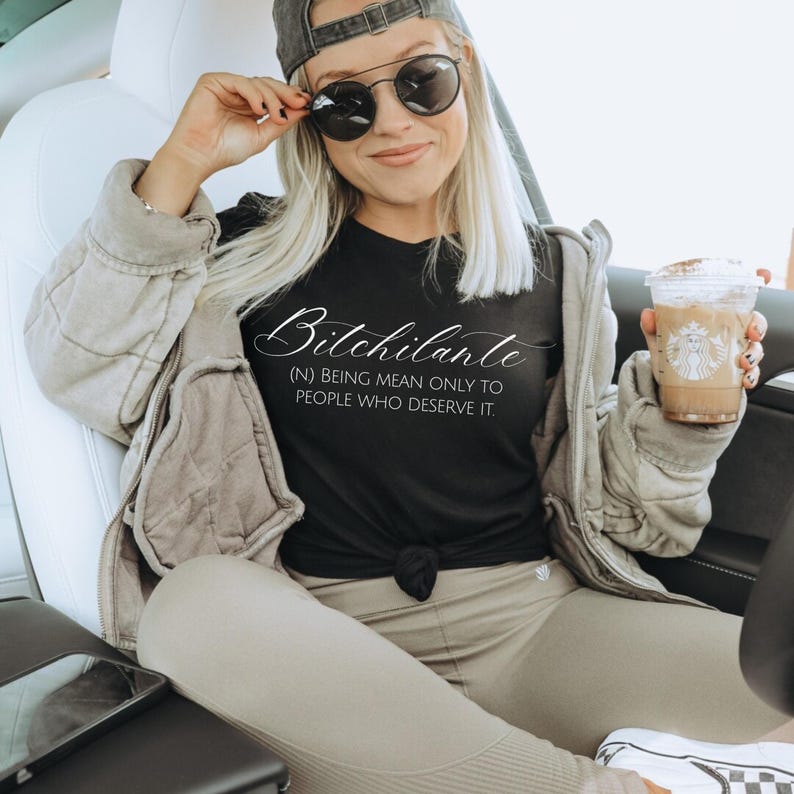 May include: Black t-shirt with the word "Bitchilante" in cursive script. Below the word is the definition: "(N) BEING MEAN ONLY TO PEOPLE WHO DESERVE IT." The person in the image is wearing sunglasses and a baseball cap.