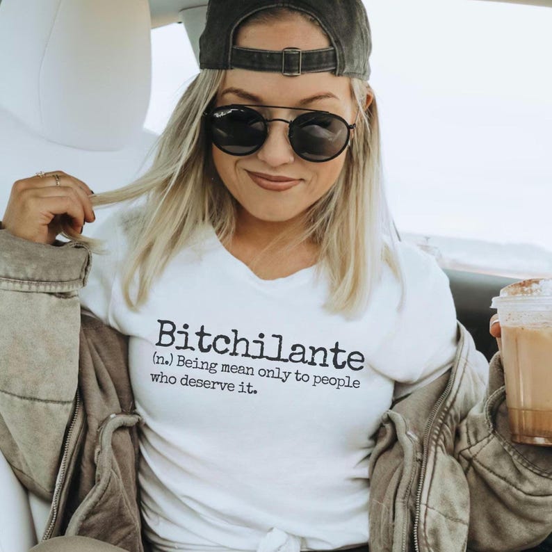 May include: White t-shirt with the word "Bitchilante" printed in black, along with a definition. The person in the image is wearing a gray baseball cap, black sunglasses, and a gray jacket. The person is holding a coffee cup.