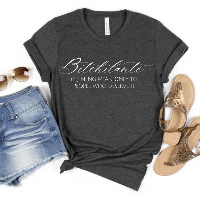 May include: Dark gray t-shirt with the word "Bitchilante" in a cursive font. Below the word is the definition: (N) BEING MEAN ONLY TO PEOPLE WHO DESERVE IT. The shirt is paired with denim shorts, sunglasses, and gold sandals.