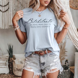 May include: Light blue t-shirt with the text "Bitchilante" in a cursive font, with the definition "(N) BEING MEAN ONLY TO PEOPLE WHO DESERVE IT." The shirt is paired with denim shorts and a brown belt.