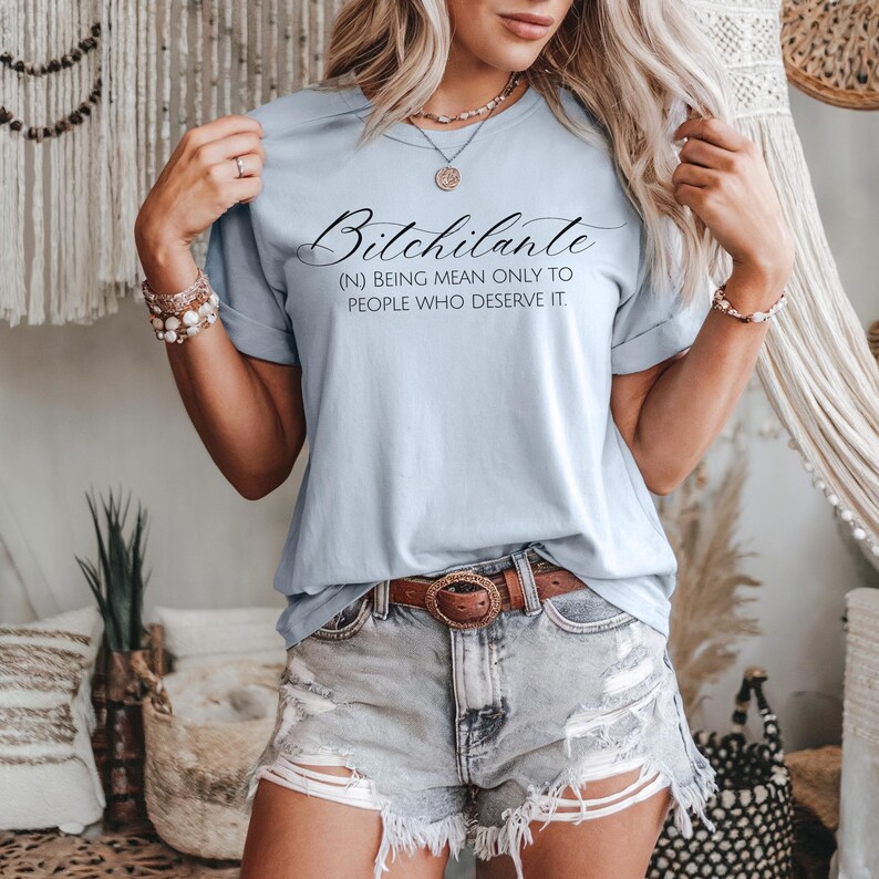 May include: Light blue t-shirt with the text "Bitchilante" in a cursive font, with the definition "(N) BEING MEAN ONLY TO PEOPLE WHO DESERVE IT." The shirt is paired with denim shorts and a brown belt.