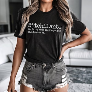 May include: Dark gray t-shirt with the word "Bitchilante" in white, along with a definition. The shirt is paired with gray denim shorts. The overall style is casual and modern.