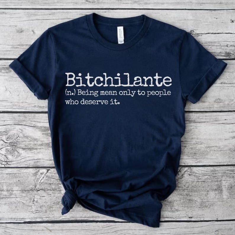 May include: Navy blue t-shirt with the white text "Bitchilante (n.) Being mean only to people who deserve it." The shirt is folded and has a tag on the collar.